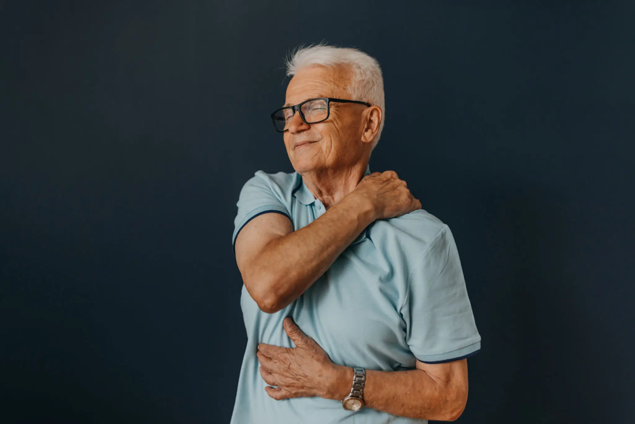 Elderly man in glasses, light blue shirt, touching his shoulder thoughtfully.