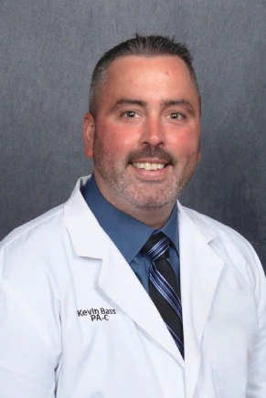 A man wearing a white doctor's coat and tie smiles at the camera.